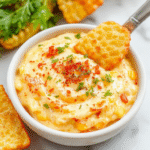A vibrant bowl of crack corn dip topped with shredded cheese and chopped herbs, surrounded by crispy tortilla chips on a rustic wooden table, with colorful ingredients in the background.