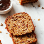 A golden slice of banana bread with a moist interior topped with banana slices and nuts, resting on a rustic wooden cutting board with a blurred background of a baking setting.